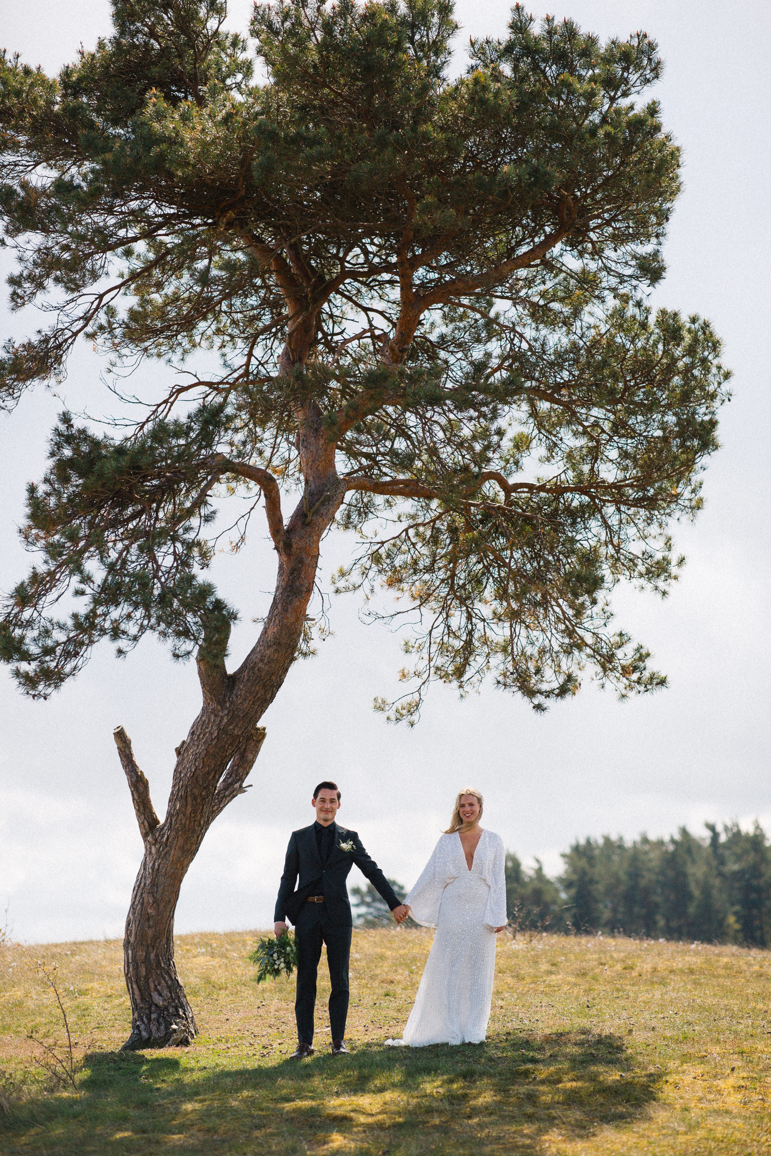 Bild i färg på brudpar som står under det ensamma trädet i Haväng under ett bröllop i Skåne. Bröllopsfotograf är Tove Lundquist. Brudklänning från Asos, brudgummens kläder kommer från Tiger of Sweden.