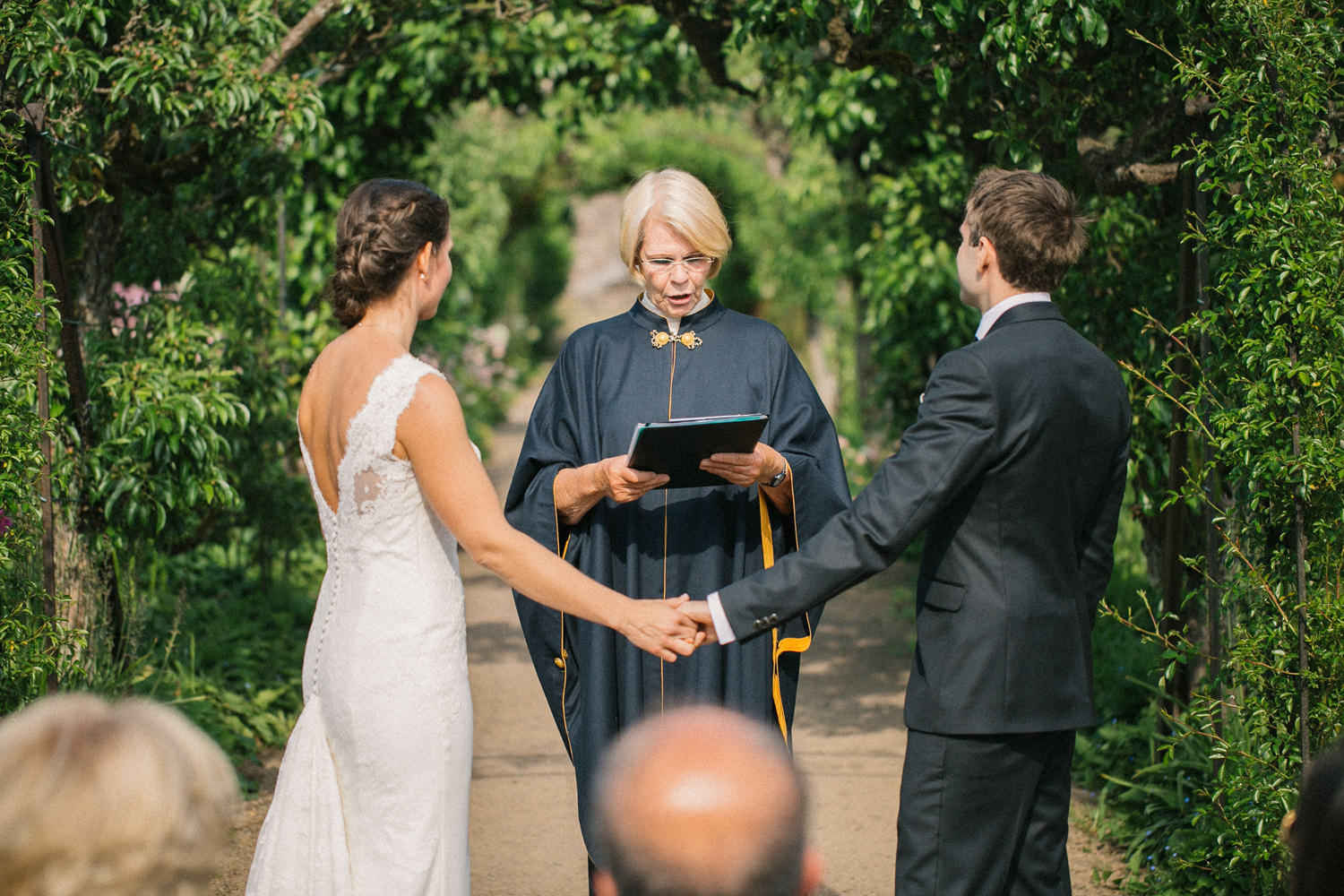 Bild i färg på brudpar med präst under en utomhusvigsel i trädgården på Sofiero Slott, Helsingborg i juni. Foto: Tove Lundquist som är bröllopsfotograf i Skåne.